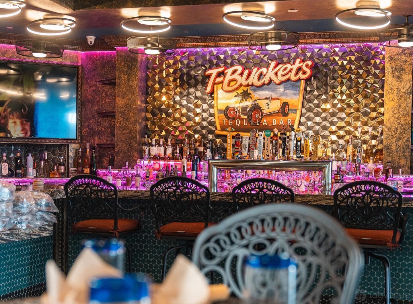 A restaurant bar area with a patterned metallic wall, neon T-Buckets sign, and a display of liquor bottles at Shooting and Social club.