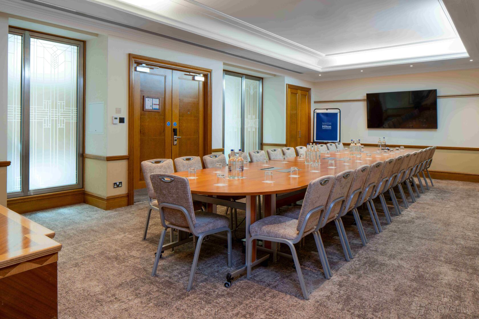 A boardroom with a long oval wooden table, upholstered chairs, wall-mounted screen, and bottled water at Hilton London Paddington.