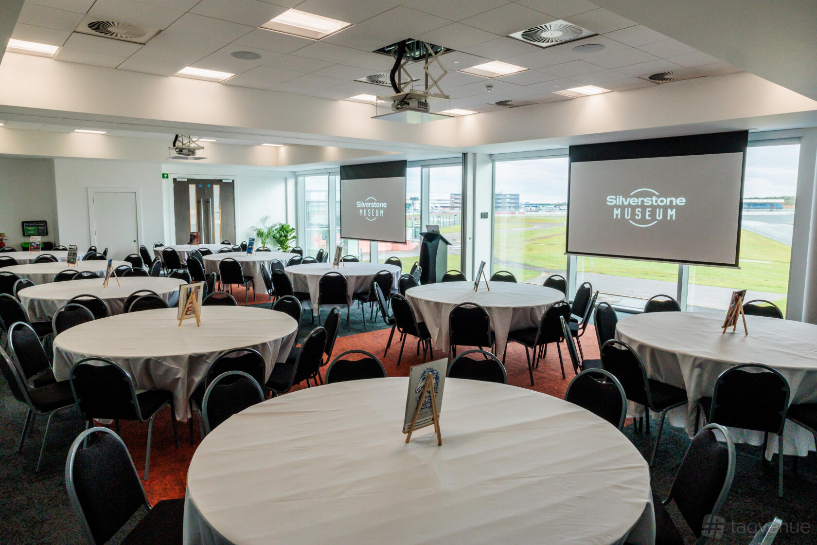 An event space in a museum with round tables covered in white linens, large windows, and projection screens at Silverstone Museum.