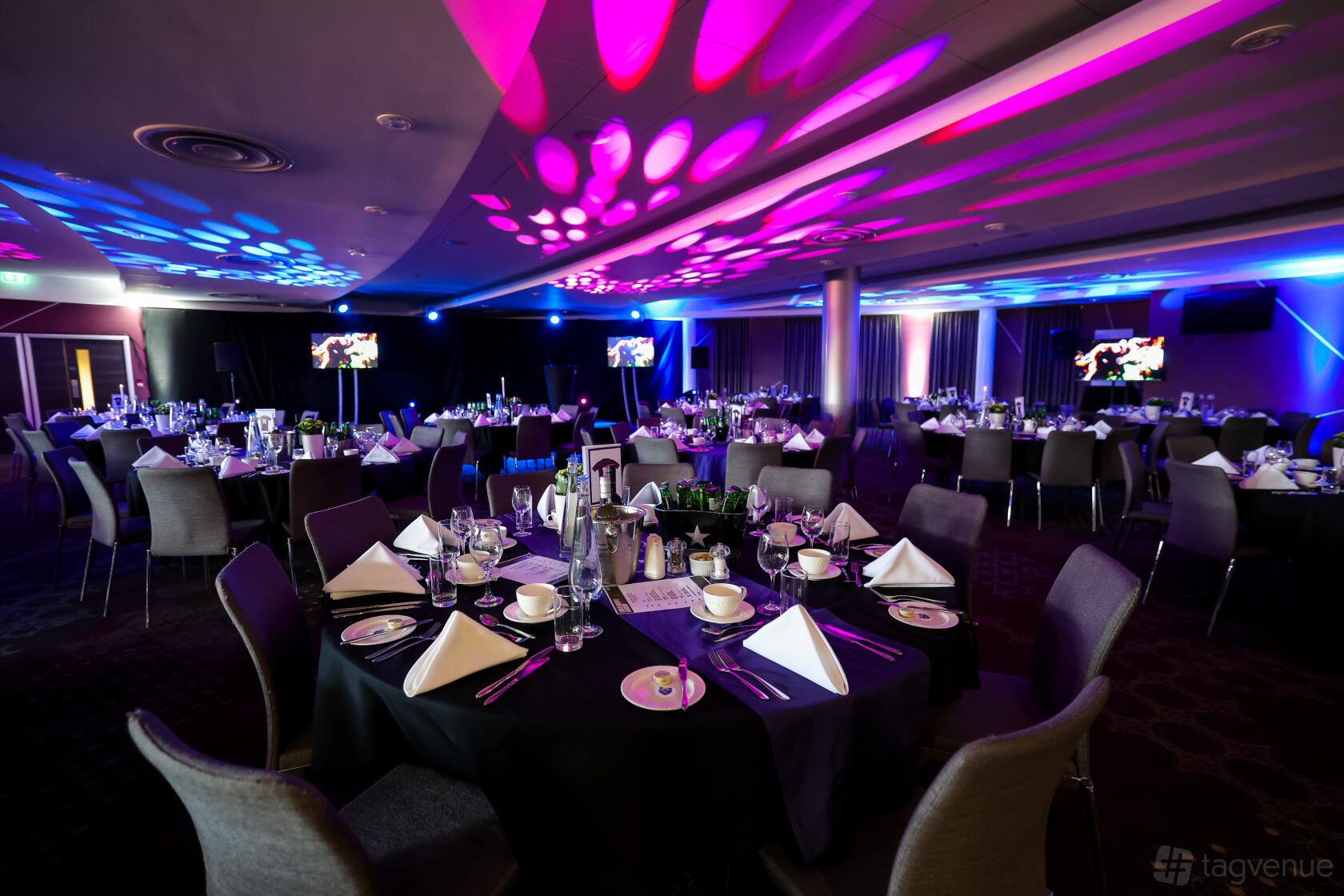 A stadium lounge with round tables set with glassware and white napkins under colorful uplighting at Ashton Gate Stadium.