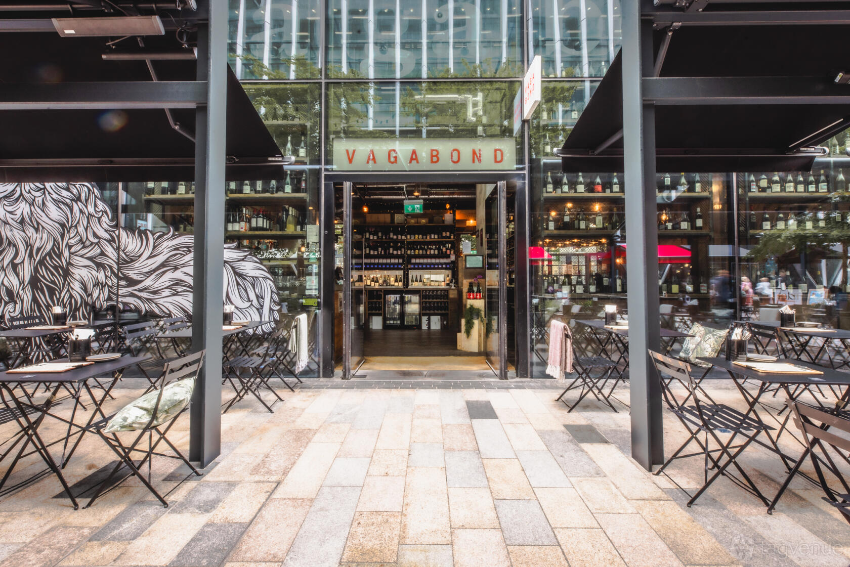 A pub with an outdoor seating area of black metal tables and chairs, glass facade, and shelves of bottles at Vagabond Victoria.