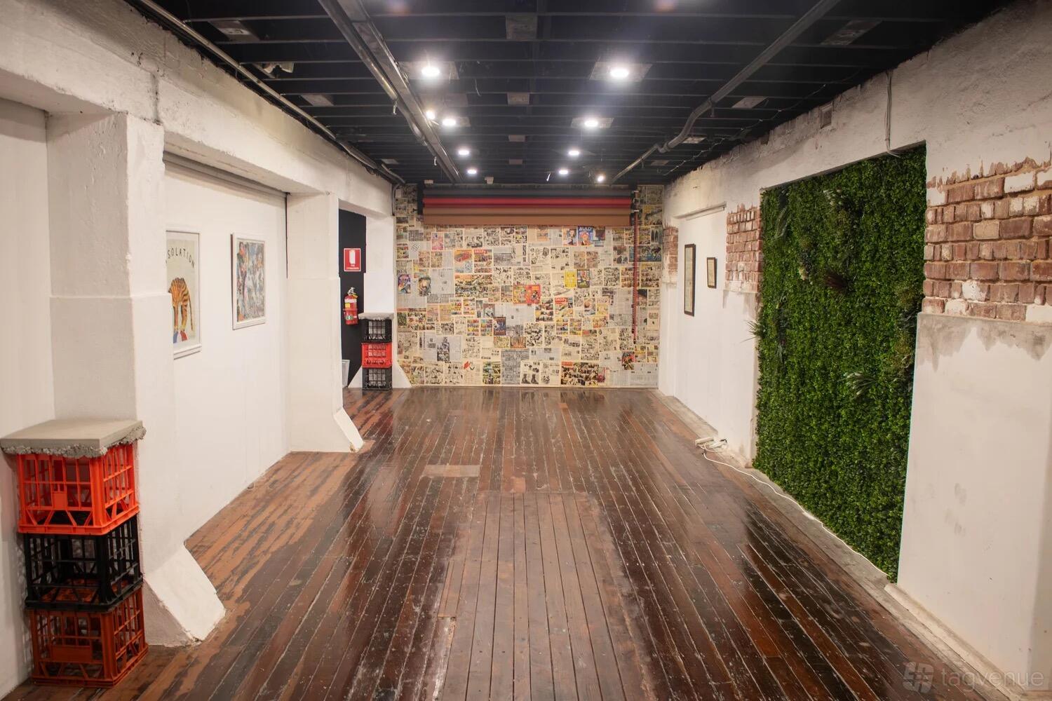 A basement event venue with polished wooden floors, exposed ceiling beams, and a collage accent wall at The Basement Gallery & Studio.