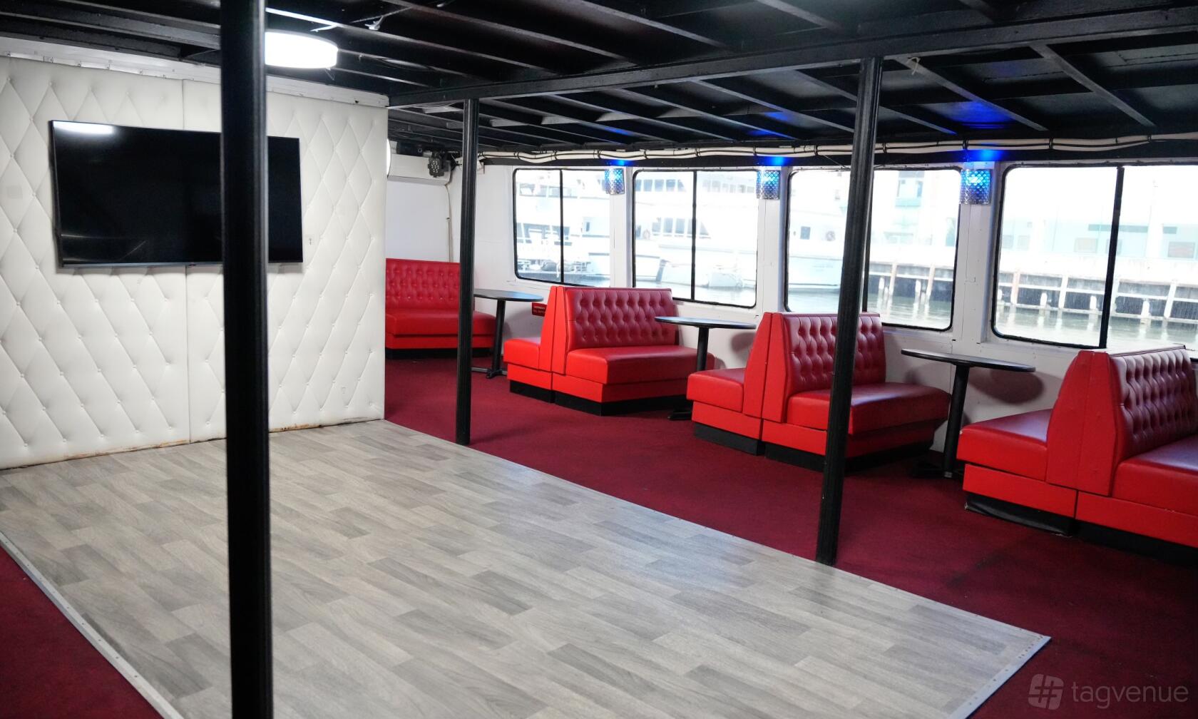 A yacht interior with red leather booths, large windows, wood flooring, and wall-mounted TV at Avalon Yacht – Skyport Marina NYC.