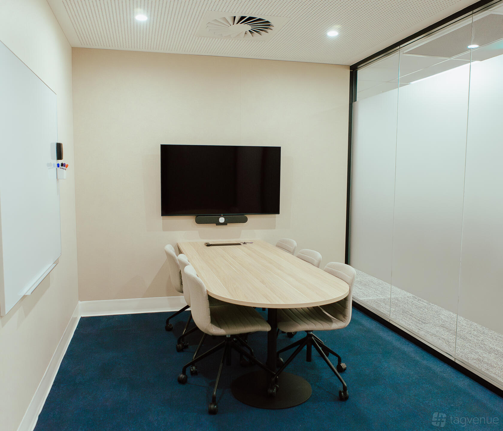 A meeting room with a wall-mounted screen, oval table, six chairs, glass wall, and whiteboard at Tank Stream Suites.