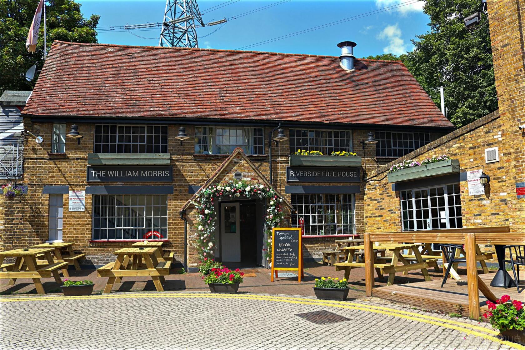 A traditional pub with a red tiled roof, outdoor wooden picnic tables, and floral decorations at The William Morris.