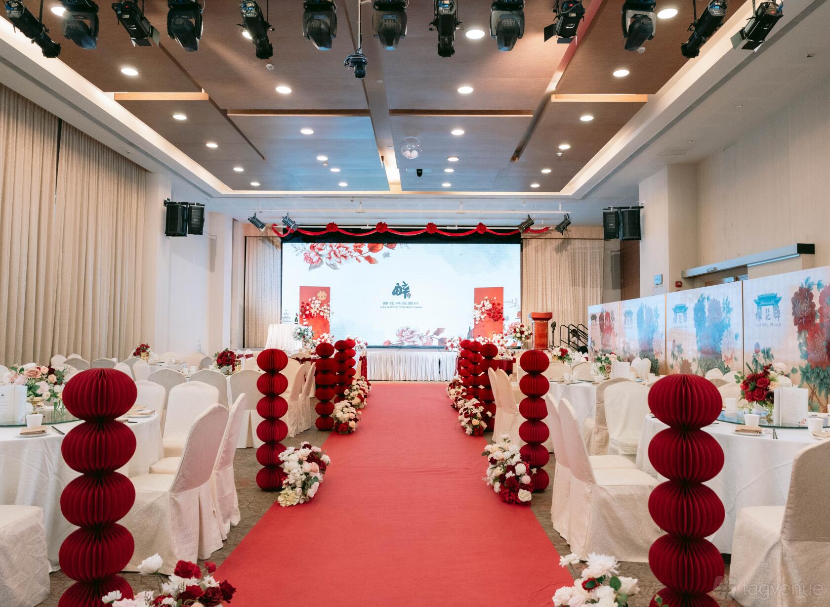 A banquet hall with red aisle carpet, white covered chairs, floral arrangements, and a decorated stage at Chui Huay Lim Teochew Cuisine.