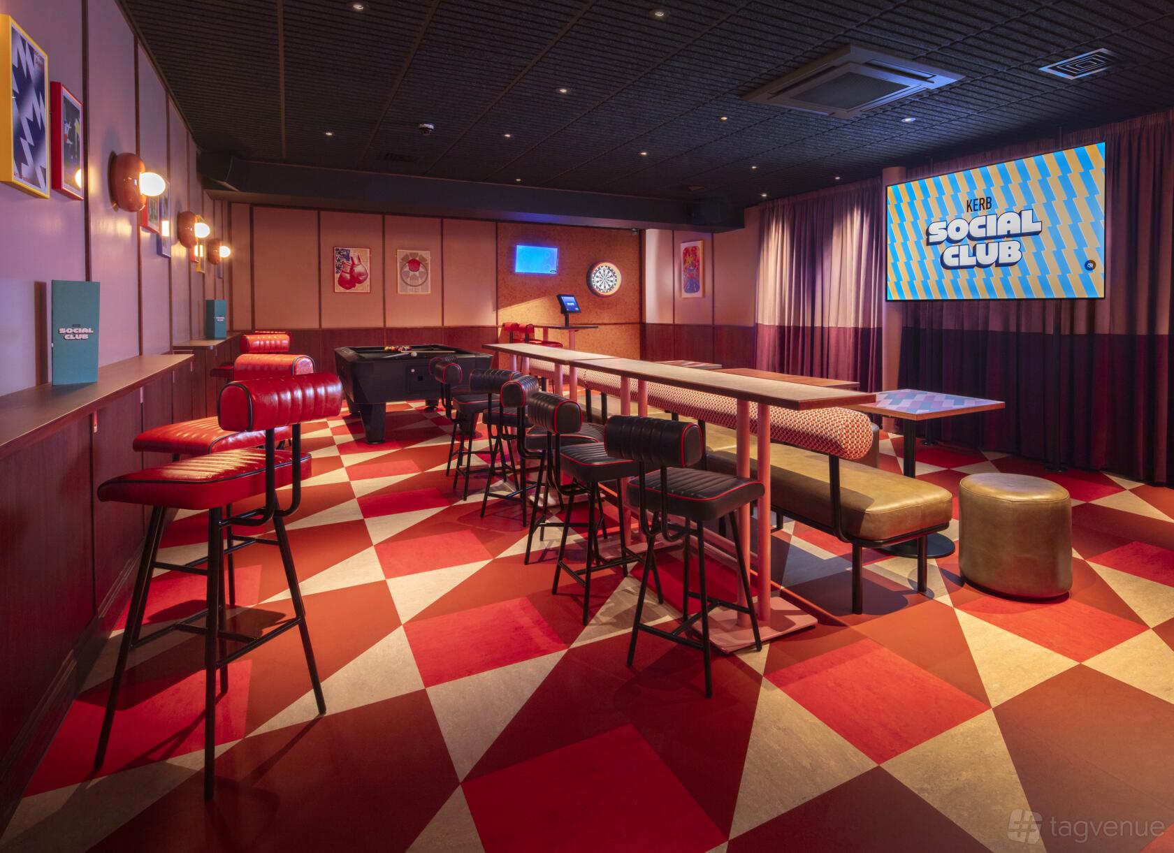An event space in a bar with checkered flooring, high tables, red bar stools, and a large screen at KERB Social Club Spitalfields.
