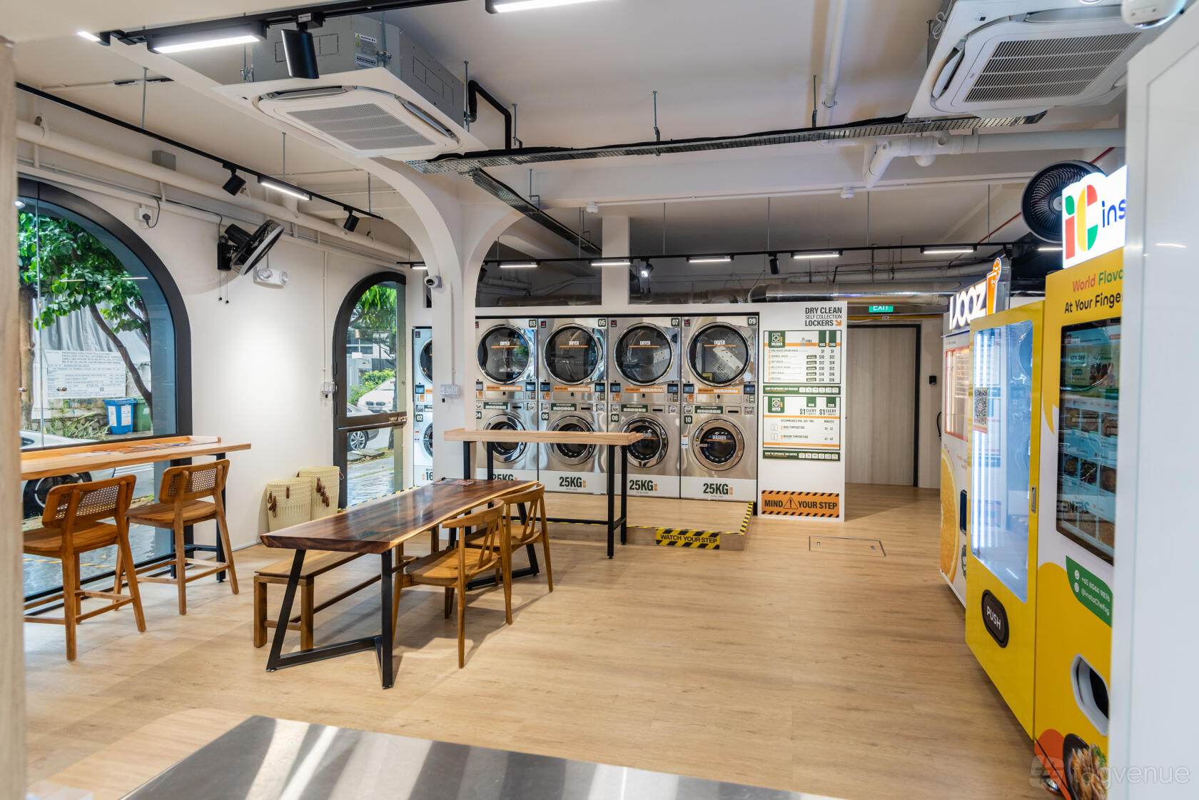 An event venue with long wooden tables, metal stools, washing machines, and large arched windows at The Good Husband Laundromat & Drycleaners.