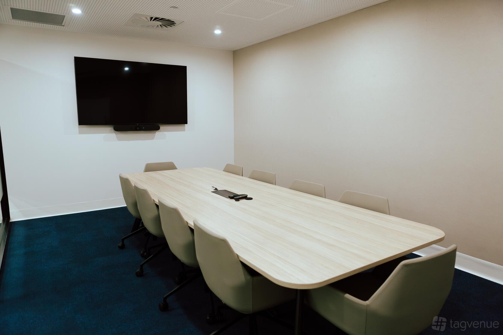 A meeting room with a long wooden table, ten beige chairs, and a wall-mounted screen at Tank Stream Suites.