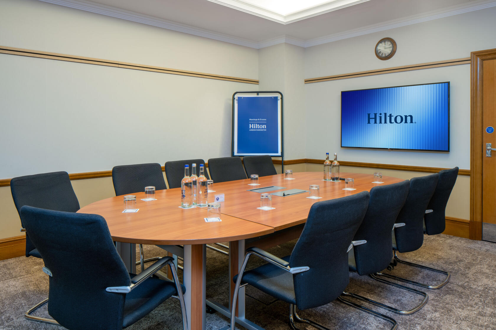 A boardroom with an oval wooden table, black chairs, wall-mounted screen, and bottled water at Hilton London Paddington.