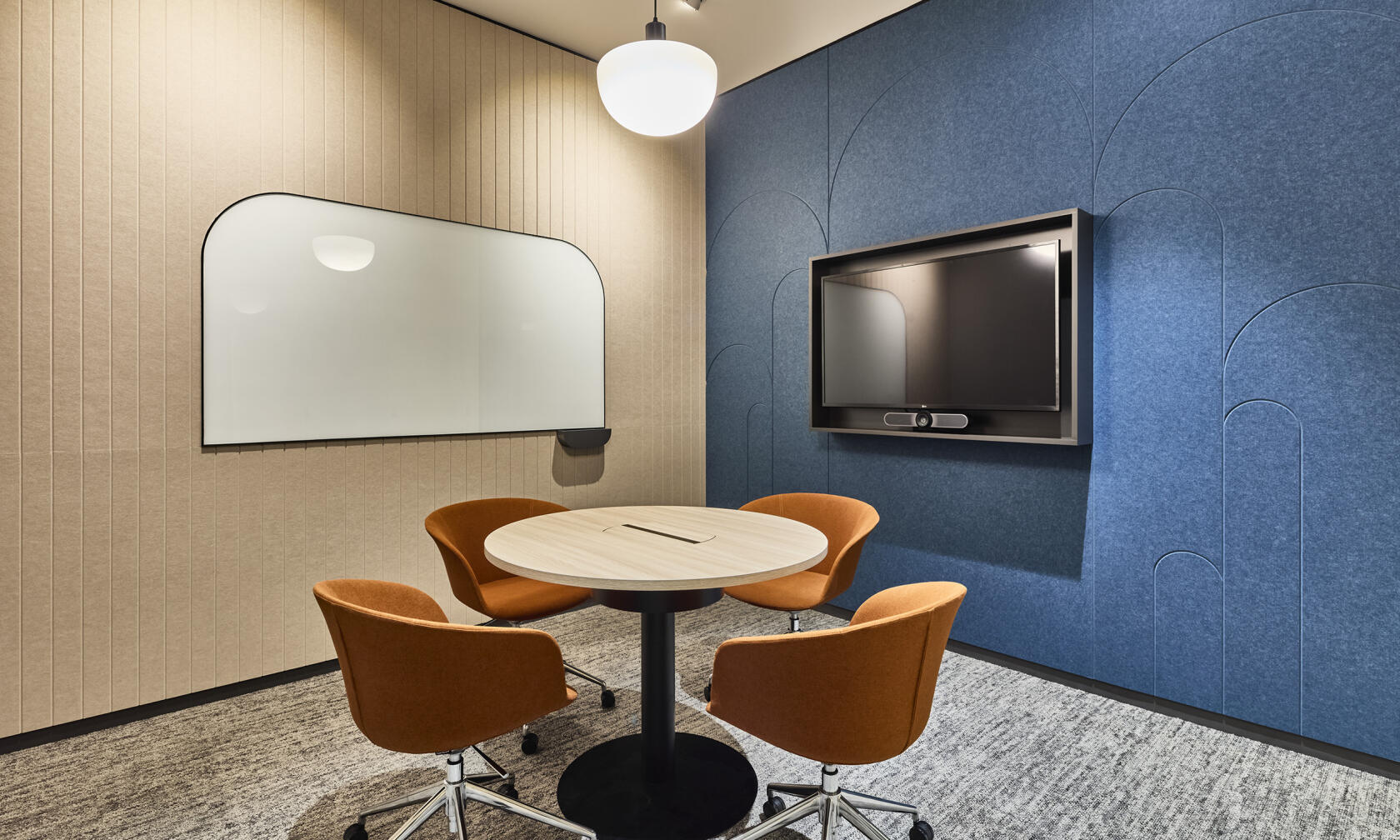 A meeting room with a round table, four orange chairs, wall-mounted whiteboard, and large monitor at SuperCo.