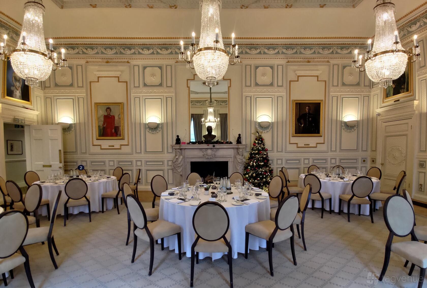 An event space in a bar with round tables set with white linens, chandeliers, portrait paintings, and a fireplace at In and Out (Naval & Military) Club.