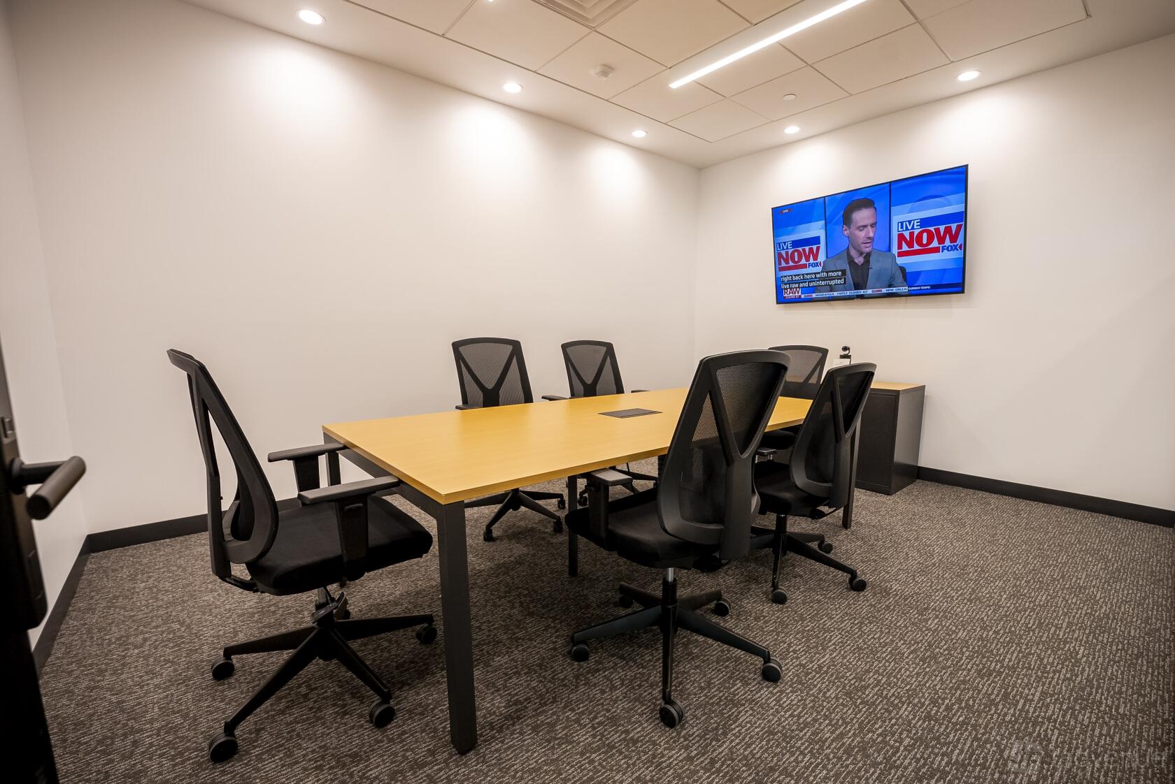 A meeting room with six black office chairs, rectangular table, wall-mounted TV, and recessed lighting at Venture X Doral.