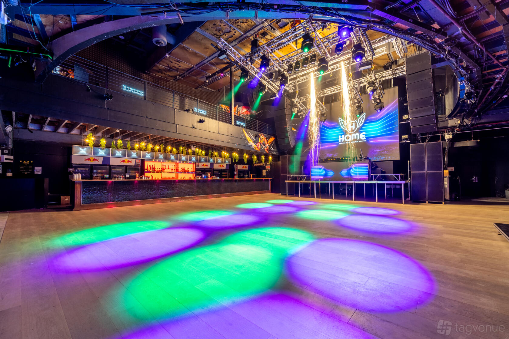 A spacious event venue with a large dance floor, colorful stage lighting, and a long bar at Home The Venue.
