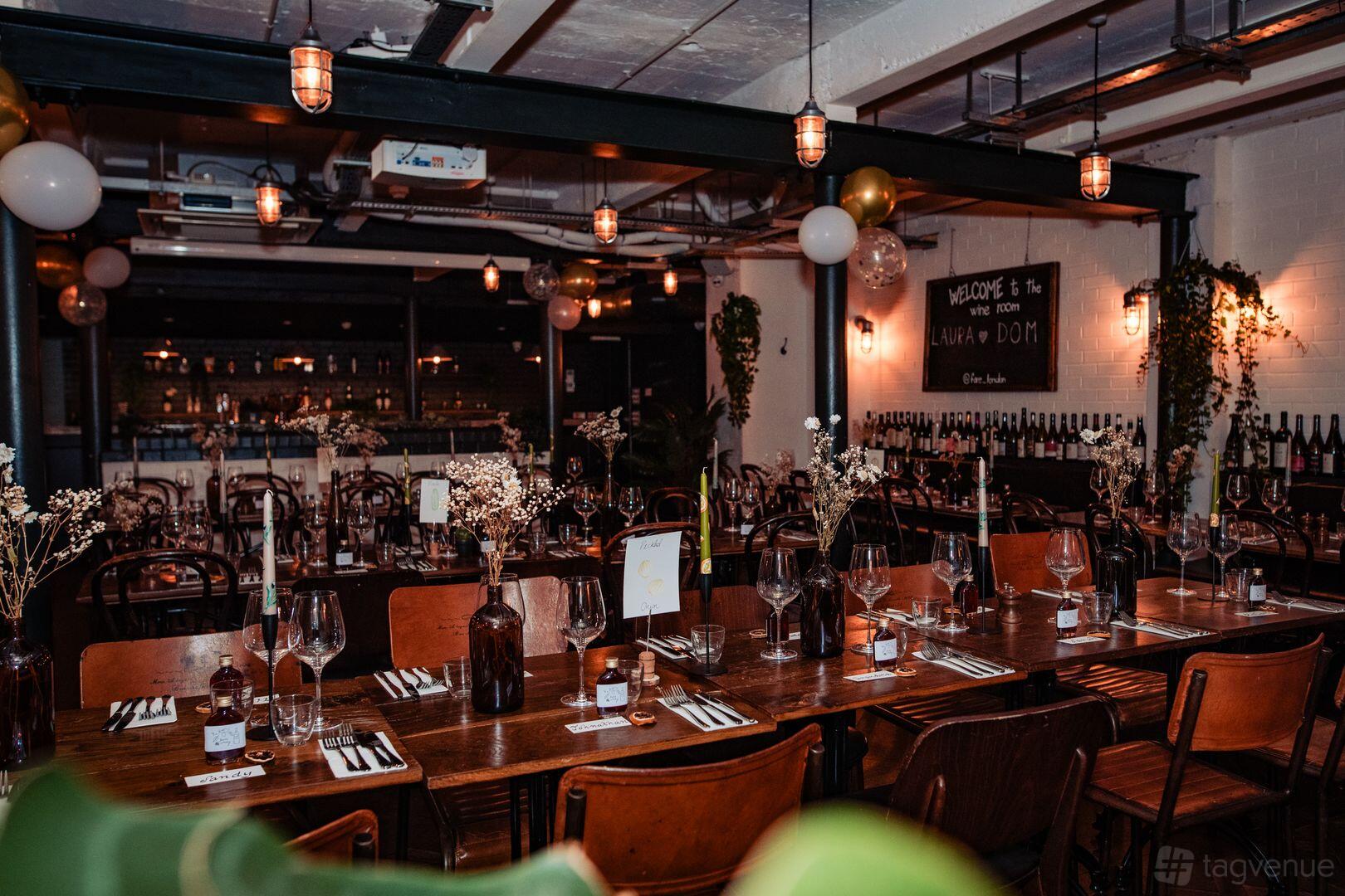 A restaurant with wooden tables set with glassware, wine bottles lining the walls, and hanging lights at Fare Restaurant + Bar.