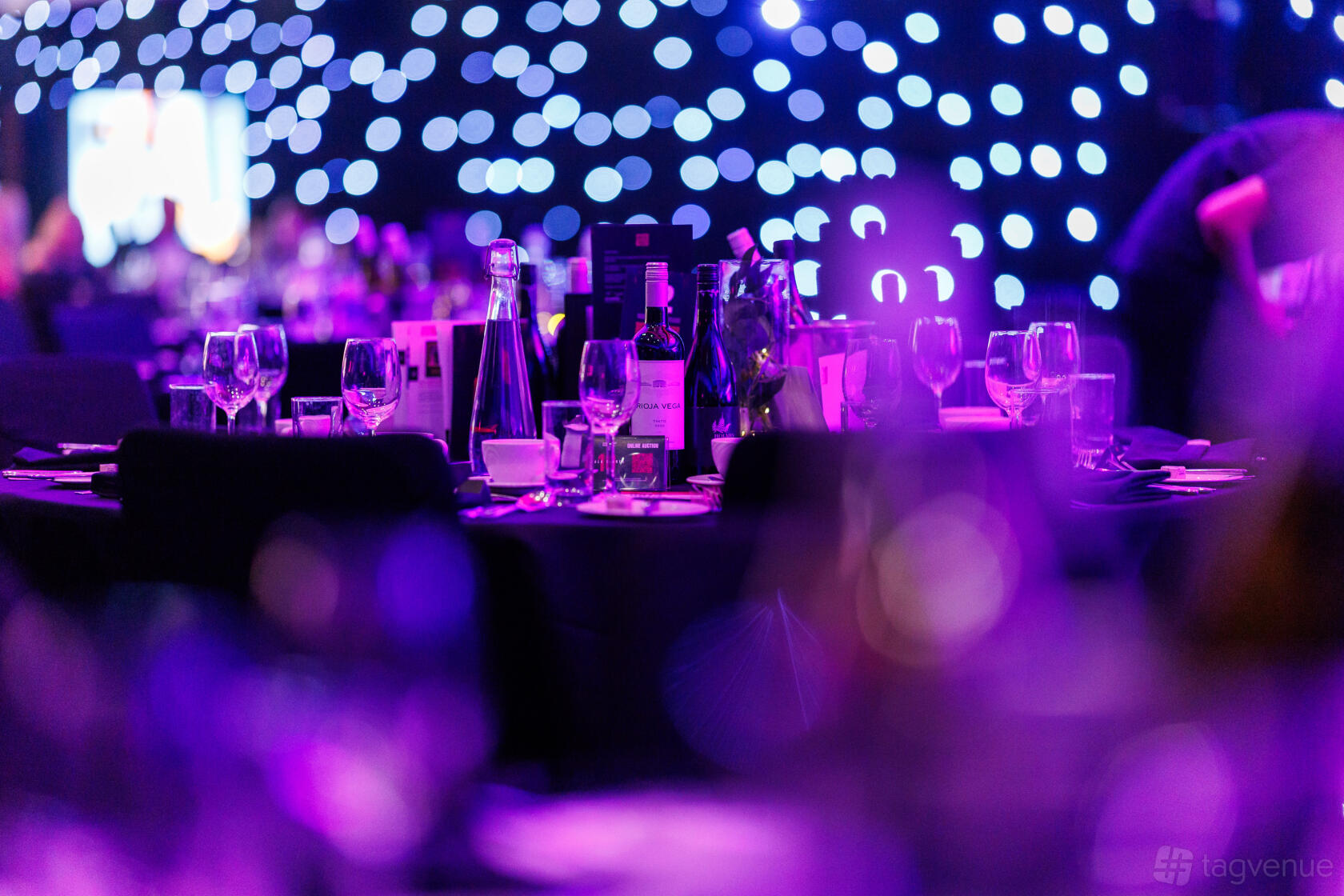 A restaurant with round tables set with glassware, bottled water, and illuminated by purple lighting at Ashton Gate Stadium.