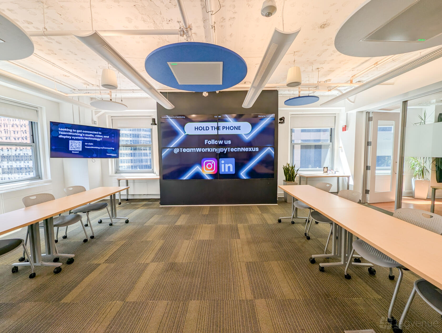 A meeting room with modular tables, ergonomic chairs, large display screens, and floor-to-ceiling windows at TeamWorking by TechNexus.