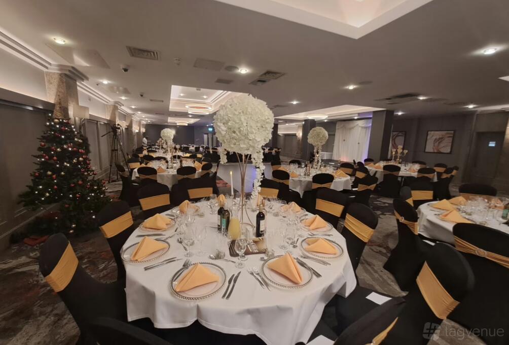A function room with round tables set with folded napkins, tall floral centerpieces, and decorated chairs at The Crown London Hotel.