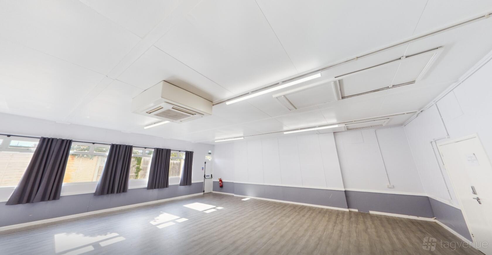 A versatile event space with large windows, dark curtains, wood flooring, and ceiling strip lights at St Patrick Church.
