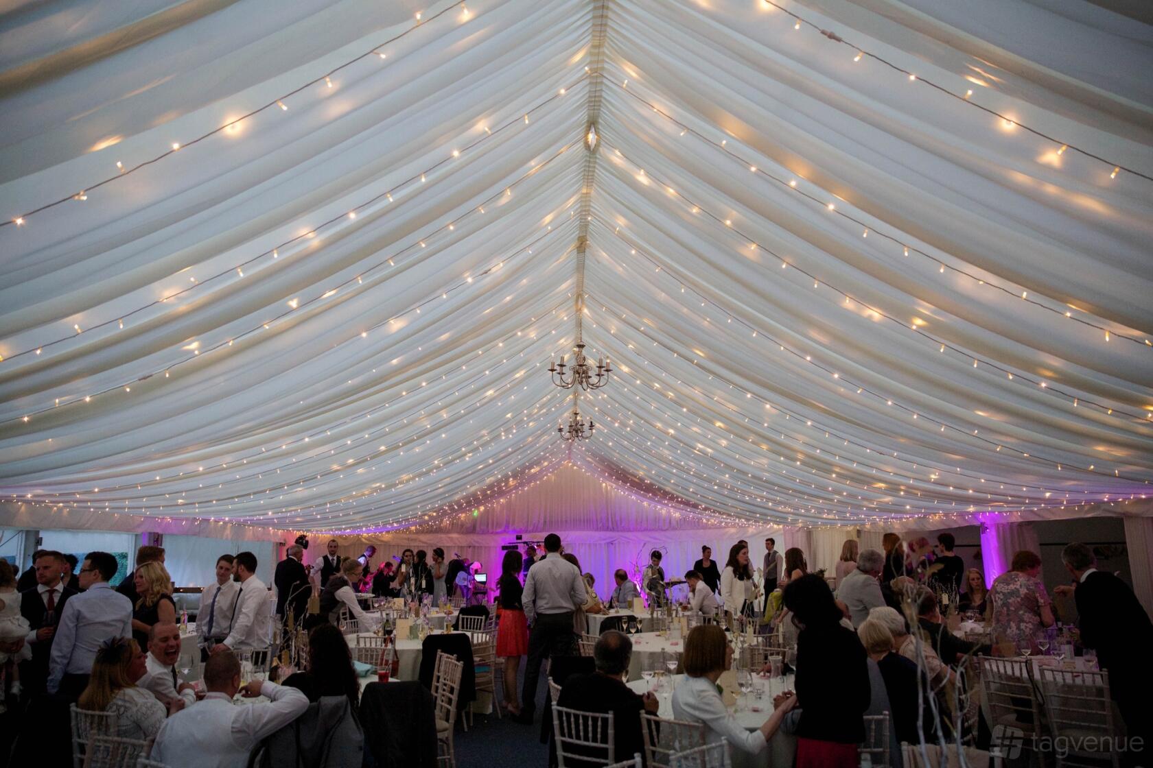 An event venue marquee with draped fabric ceiling, chandeliers, fairy lights, and round tables at Ravens Ait.