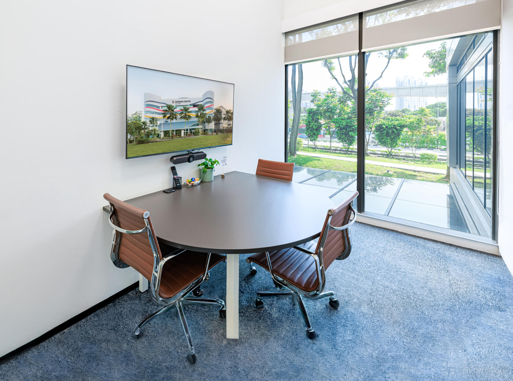 A meeting room with a round table, four brown chairs, large windows, and a wall-mounted display at German Centre Singapore.