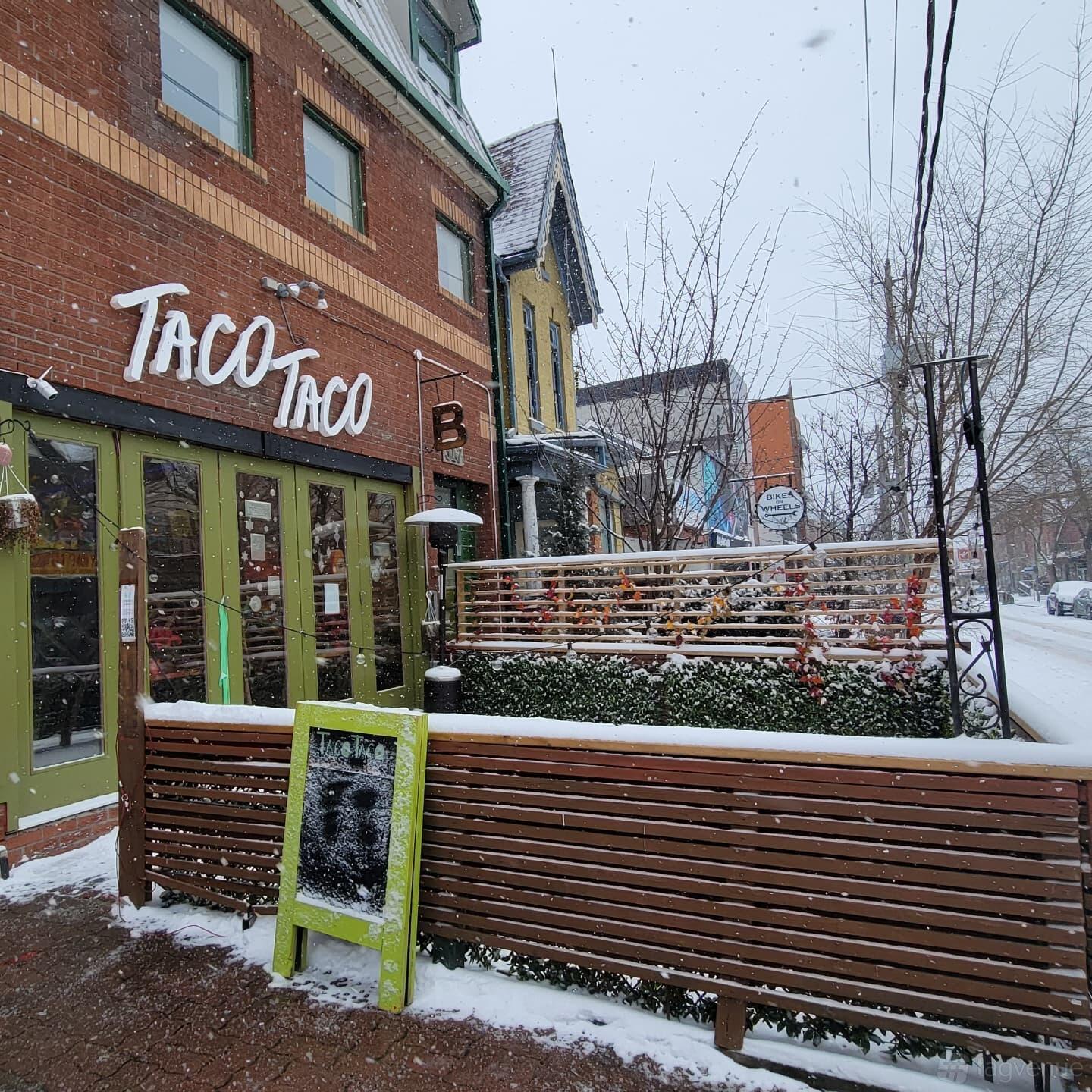 A restaurant exterior with green double doors, a wooden patio, and snow-covered benches at TacoTaco.