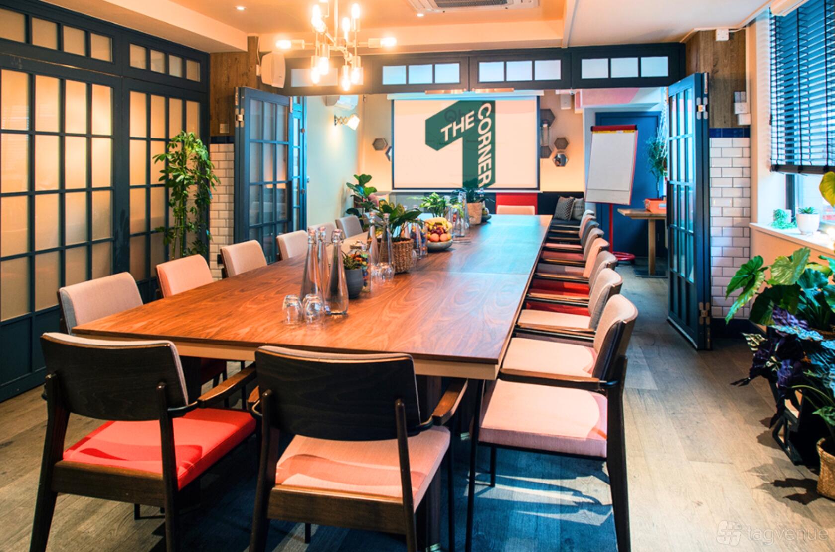 A conference room with a long wooden table, cushioned chairs, projector screen, and indoor plants at The Corner London City.