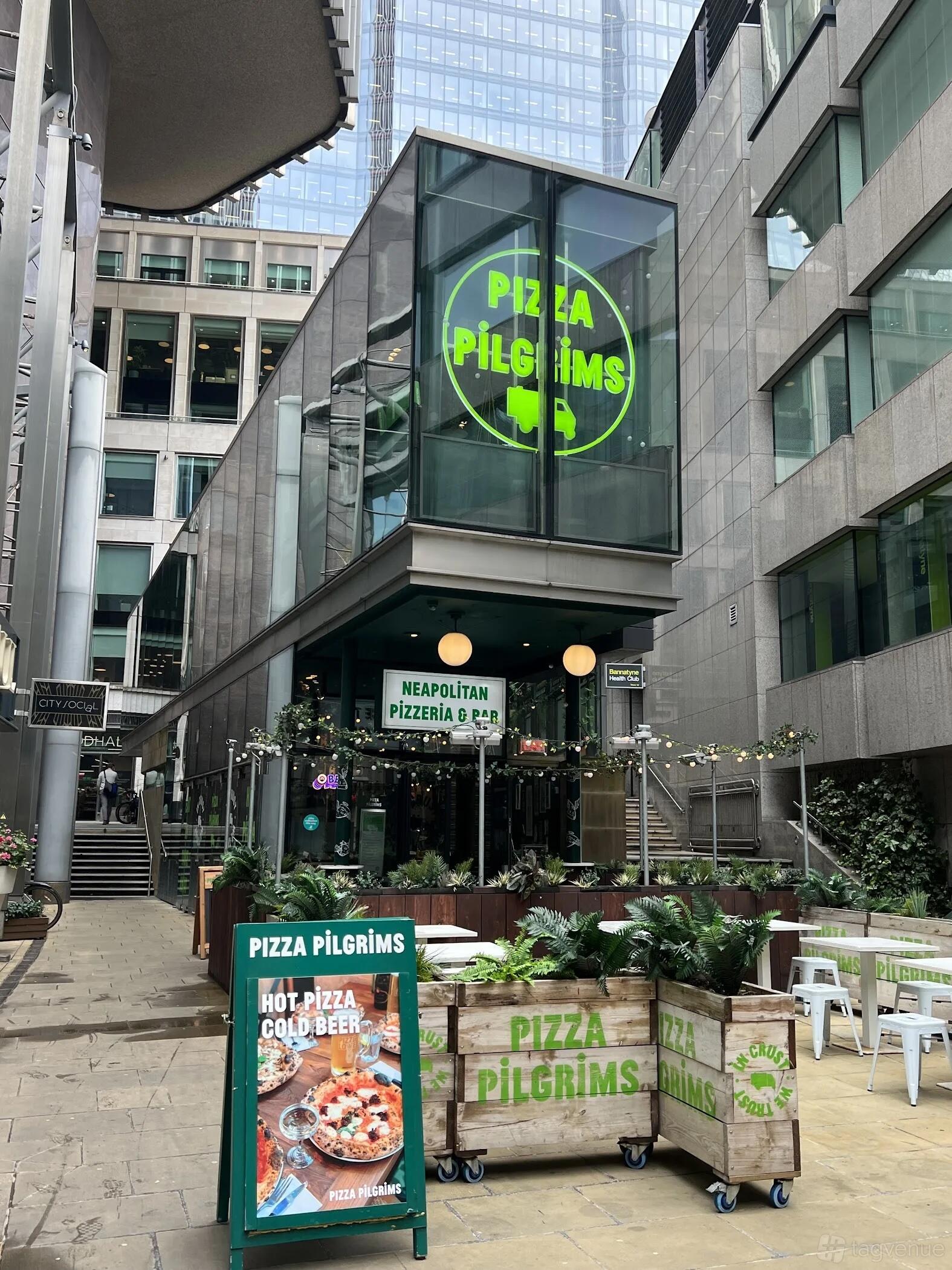 A restaurant with outdoor seating, green signage, string lights, and potted plants at Pizza Pilgrims Liverpool Street.
