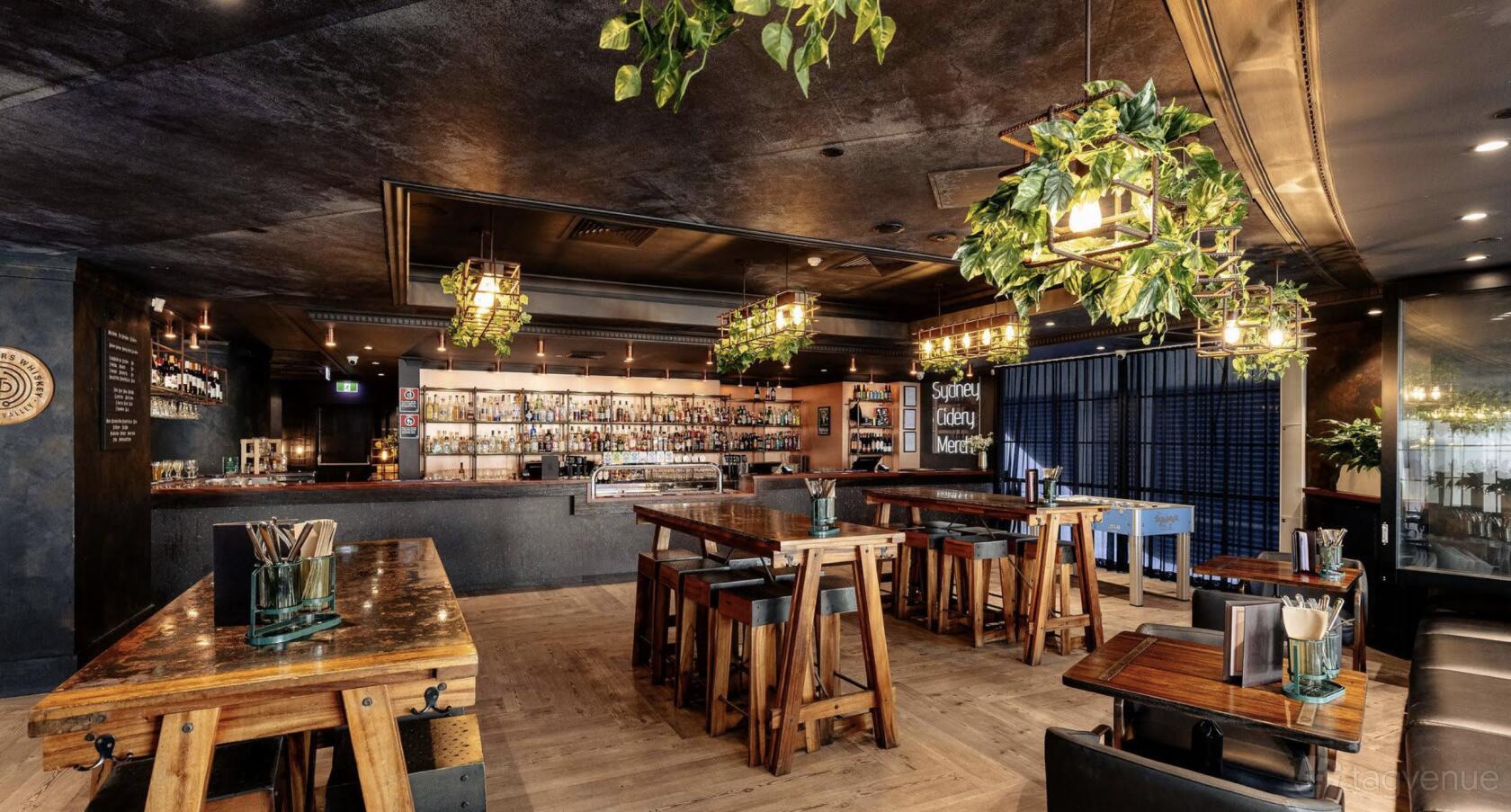A bar interior with wooden tables, hanging greenery, geometric pendant lights, and a stocked bar at Sydney Cidery.
