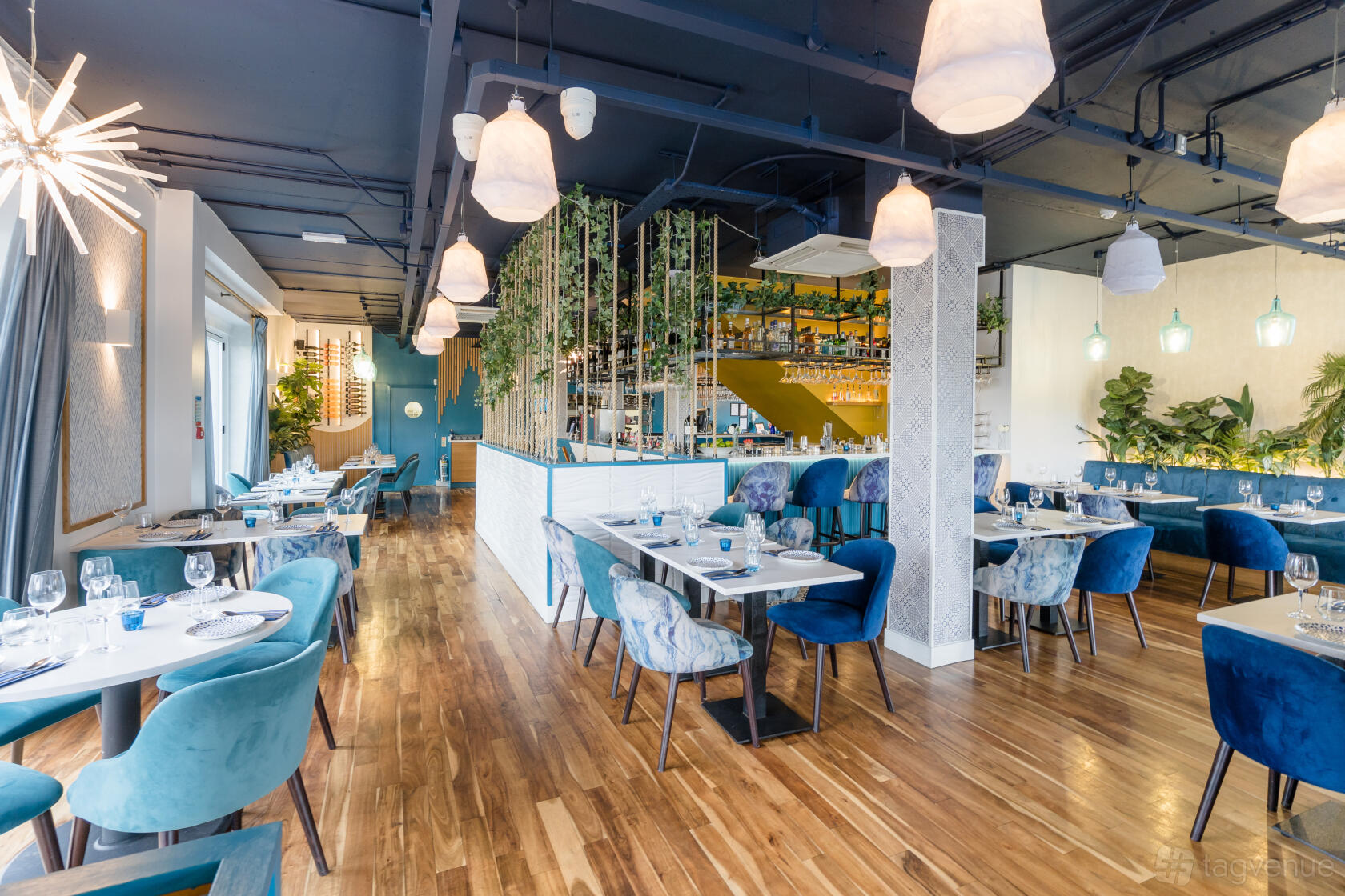 A restaurant with blue velvet chairs, wooden floors, hanging pendant lights, and greenery at El Cerdo Bourne End.