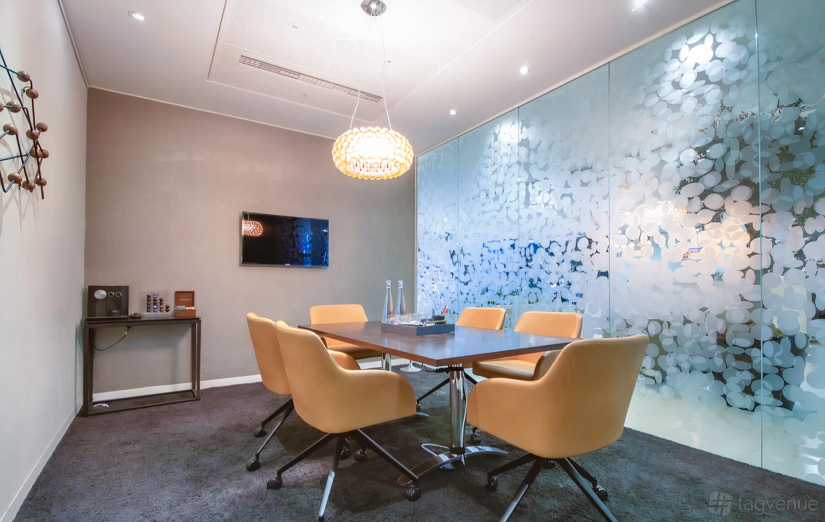 A boardroom with a rectangular table, six tan chairs, a flat-screen TV, and frosted glass walls at Regus – Belfast City Centre.