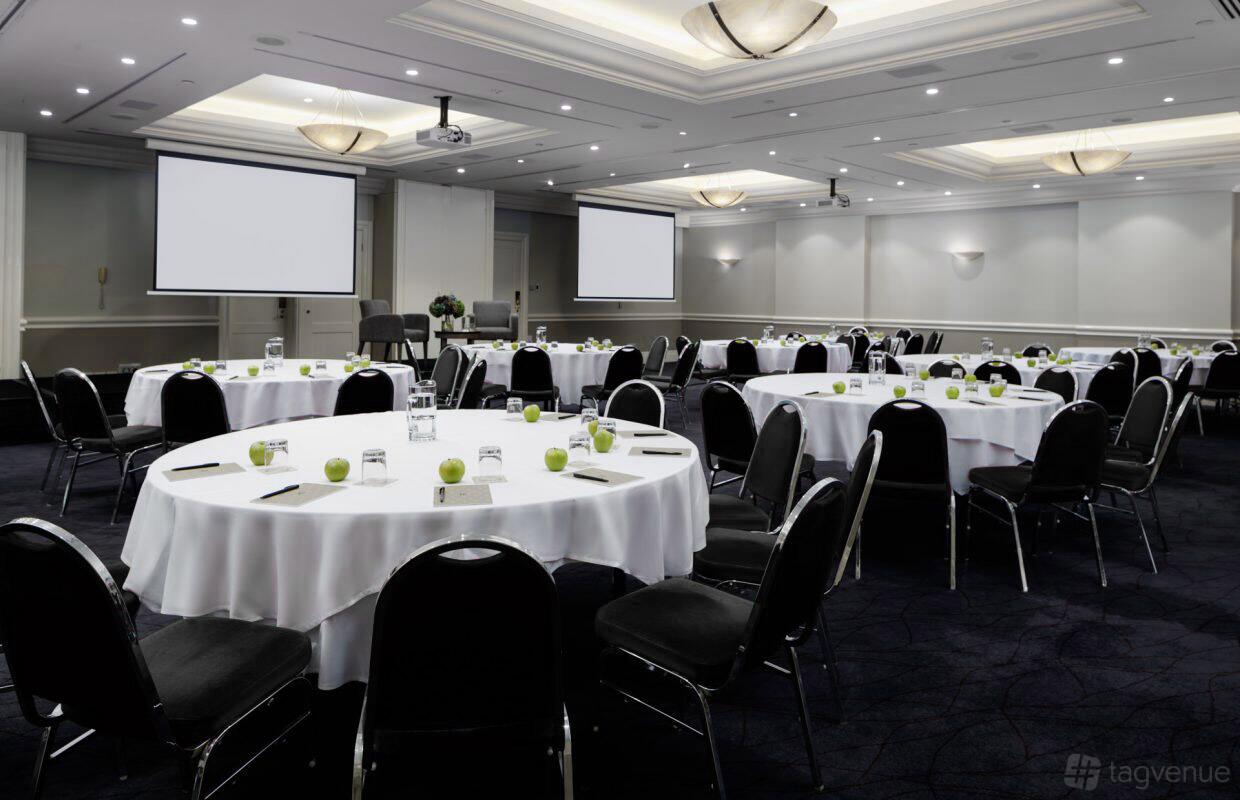 An event space with round tables covered in white linens, black chairs, and dual projection screens at Rydges Newcastle.