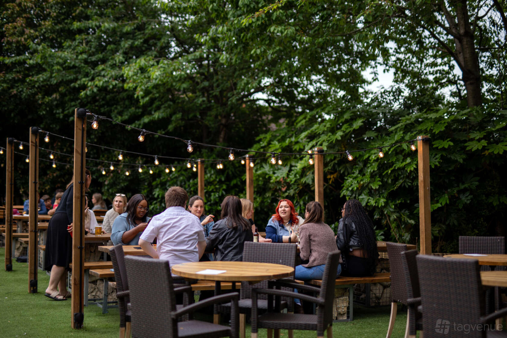 A pub garden with wooden benches, overhead string lights, and lush greenery at The Leather Bottle.