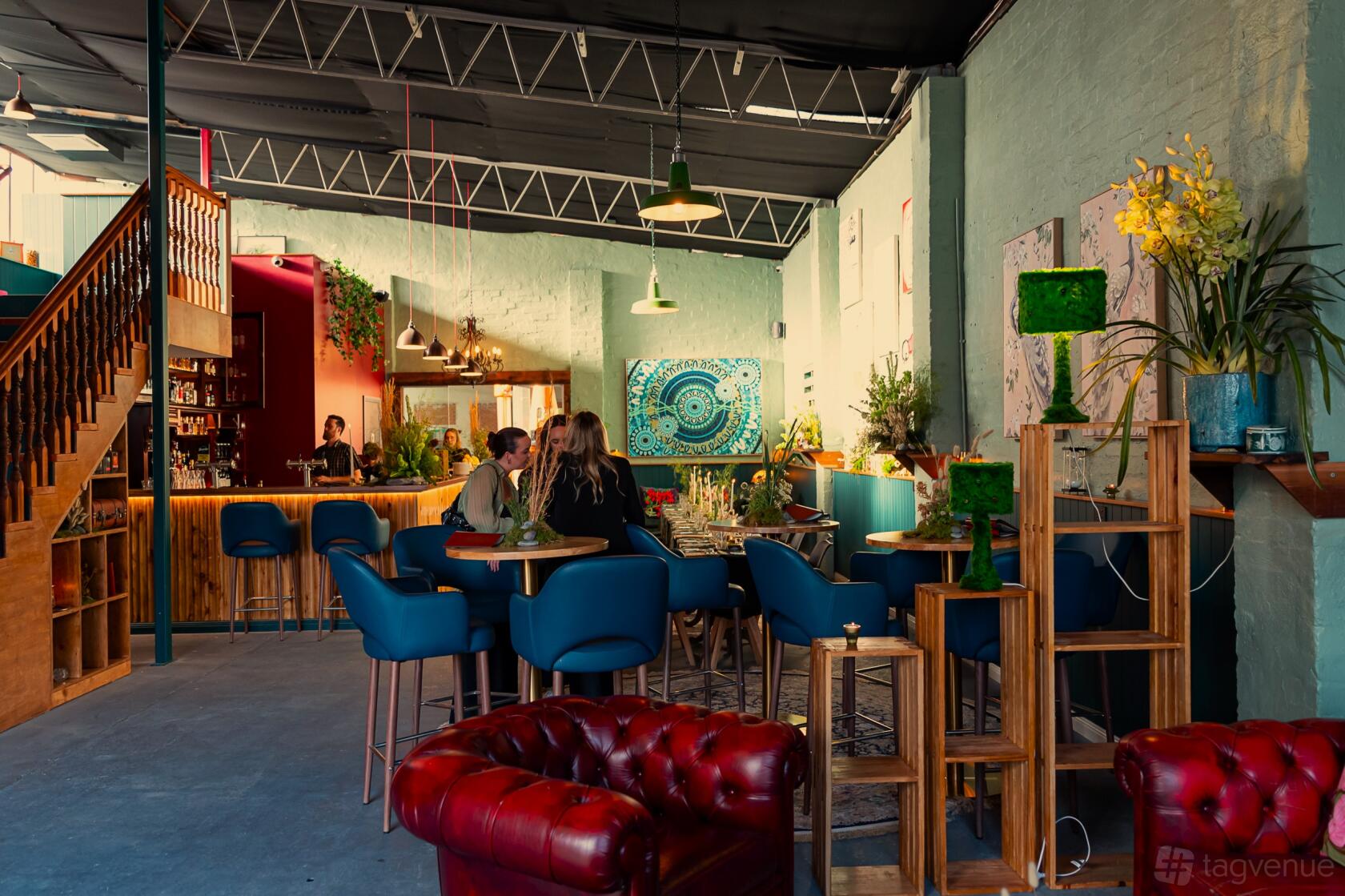 An event space in a bar with blue upholstered chairs, a wood bar, wall art, and greenery at Goodradigbee Distillers.