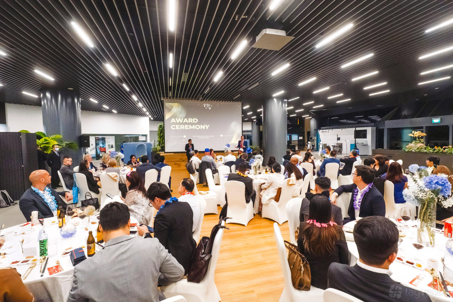 An event space with round tables draped in white linens, stage, and projection screen at German Centre Singapore.