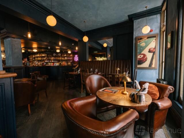 A bar in a pub with leather armchairs, round wooden tables, and pendant lights at Newhall Street Tavern.
