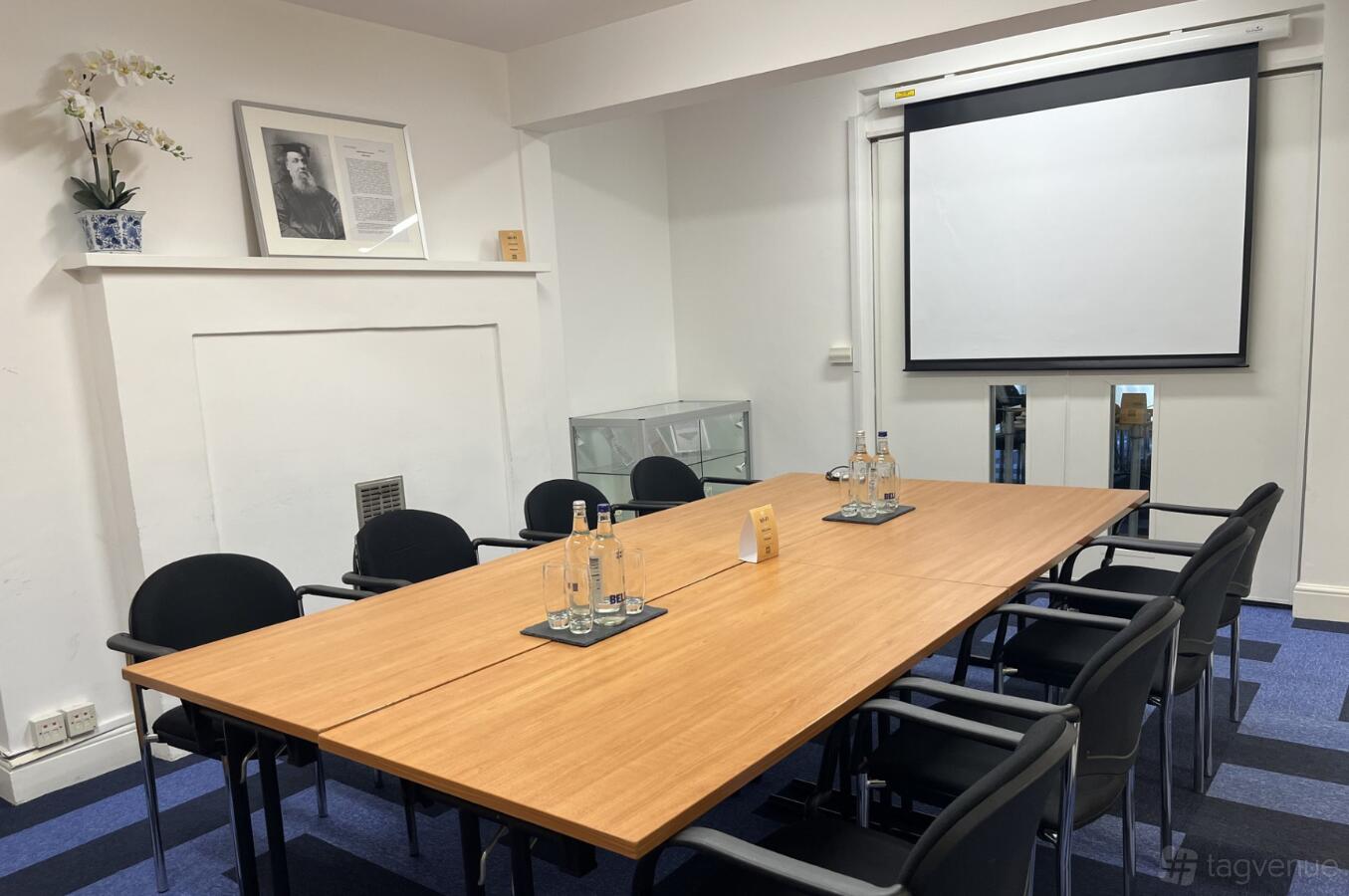 A meeting room with a rectangular table, black chairs, a wall-mounted screen, and water bottles at De Morgan House.