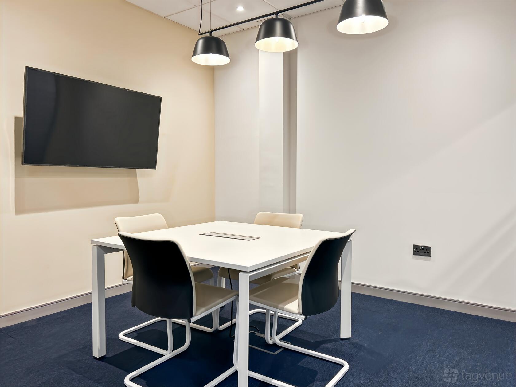 A meeting room with a square white table, four chairs, wall-mounted screen, and pendant lighting at Regus - Weavers Court.