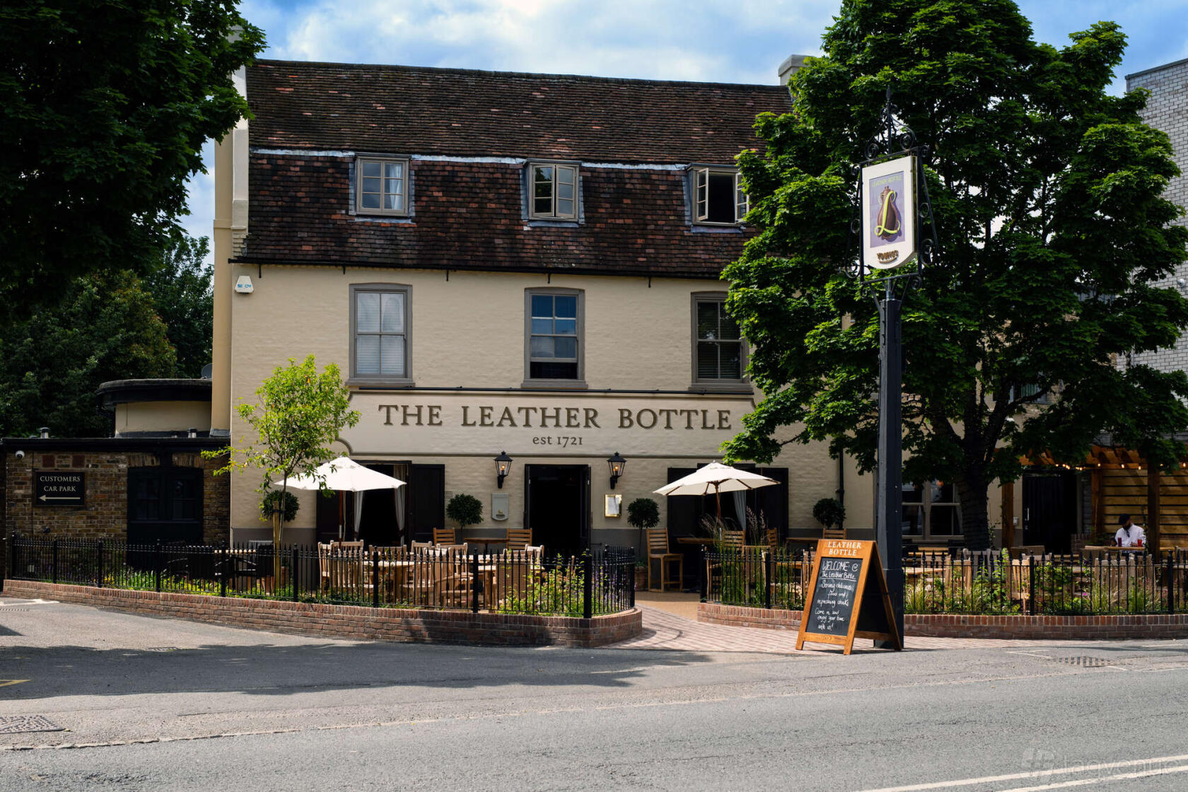 A pub with outdoor seating, wooden tables and chairs, and large trees at The Leather Bottle.