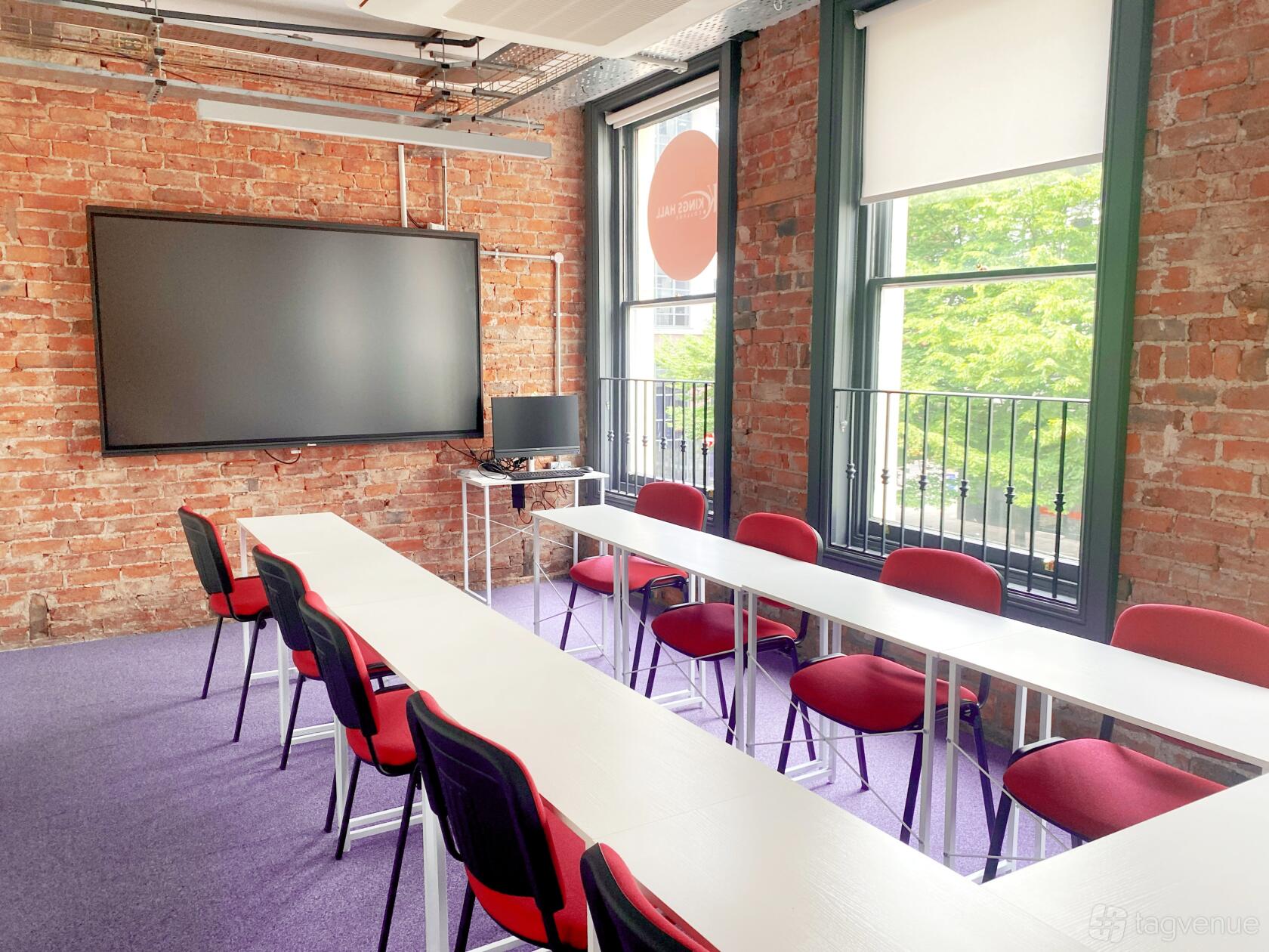 A classroom with exposed brick walls, large windows, and U-shaped white tables with red chairs at Kings Hall College.