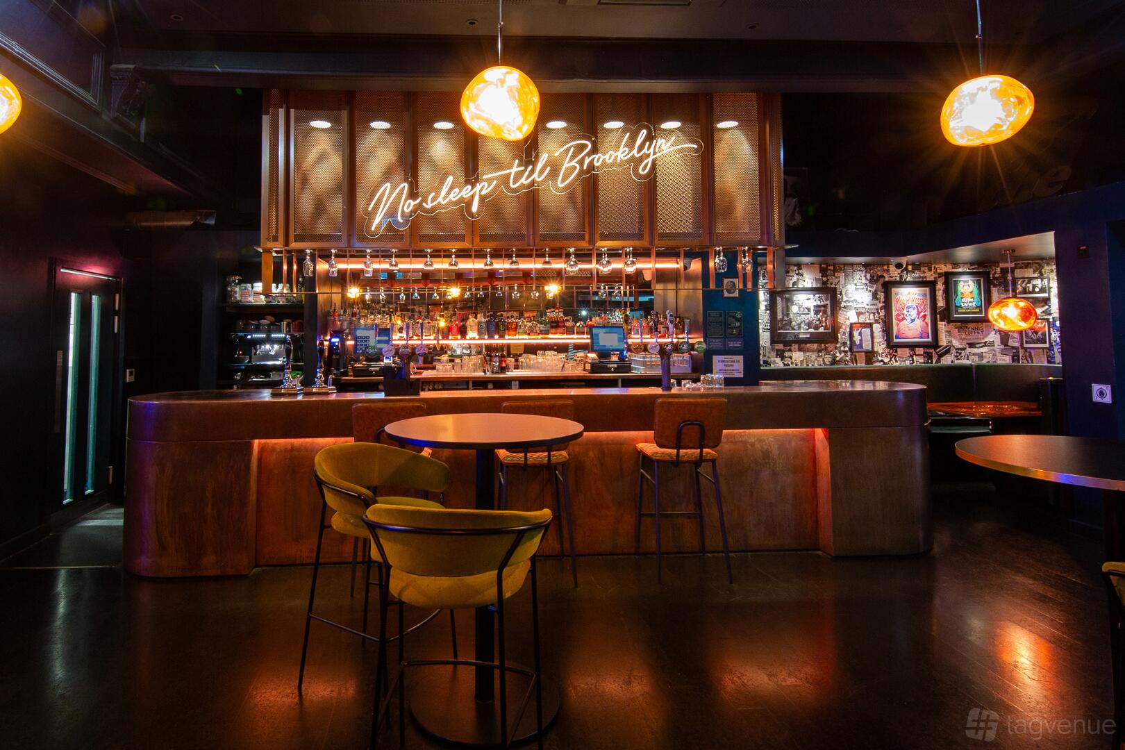 A live music venue bar with a copper counter, neon sign, and high tables with yellow stools at Band on the Wall.