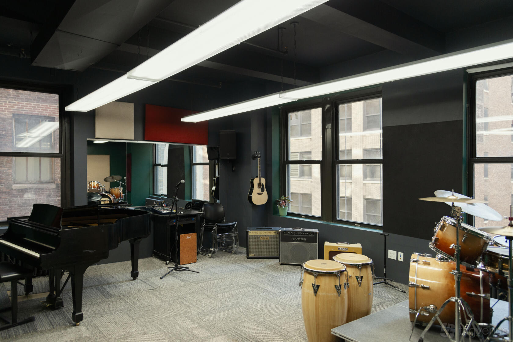 A piano room with a grand piano, drum set, congas, amplifiers, and large windows at Michiko Rehearsal Studios.