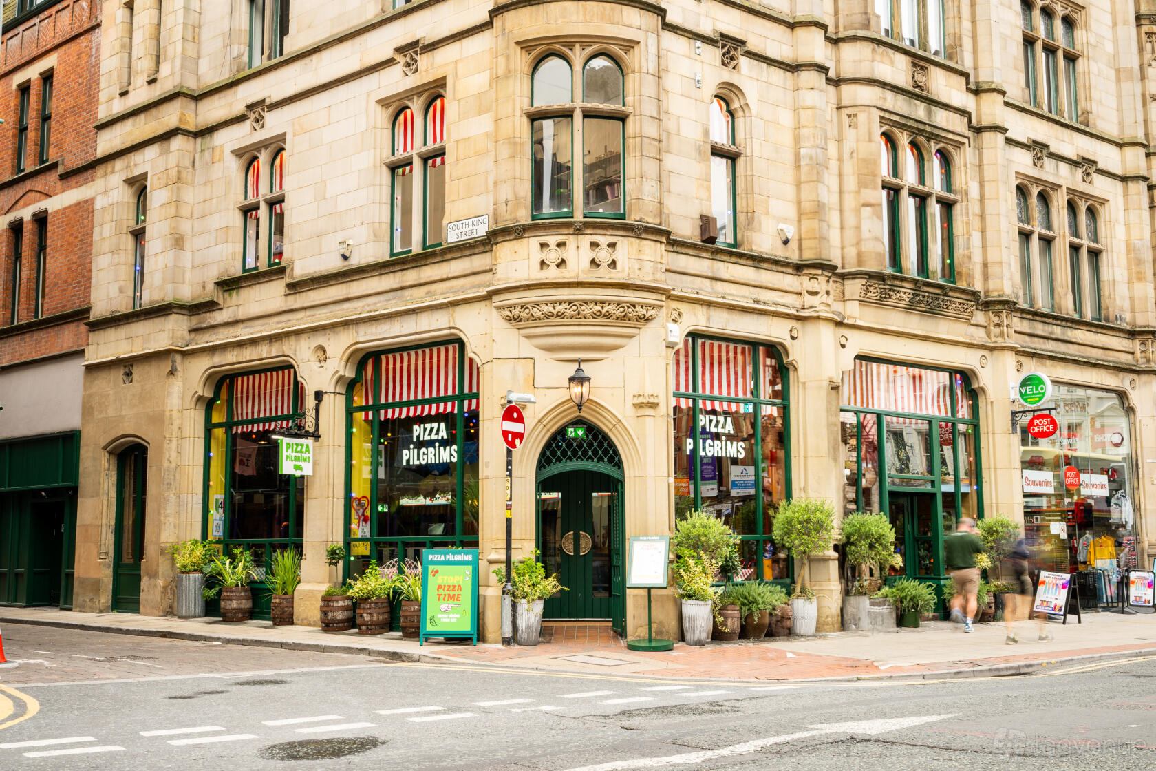 A restaurant with ornate stone façade, large arched windows, and potted plants at Pizza Pilgrims Manchester.