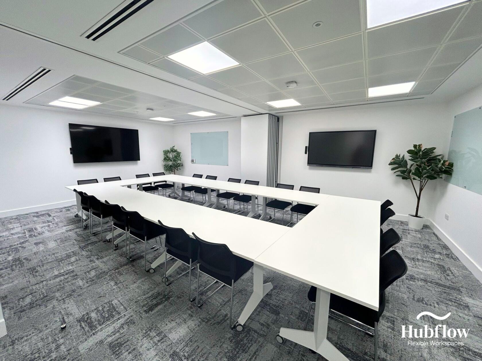 A meeting room with U-shaped white tables, black chairs, wall-mounted screens, and glass boards at Hubflow Ltd.
