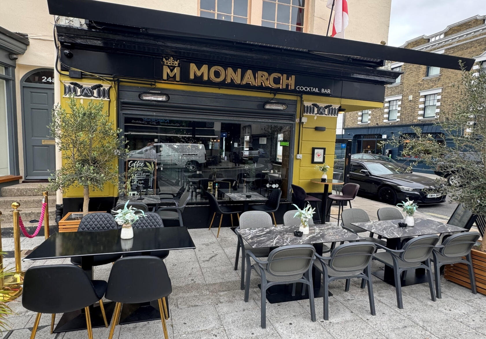 An outdoor bar area with black marble tables, potted olive trees, and cushioned bench seating at Monarch Cocktail Bar.