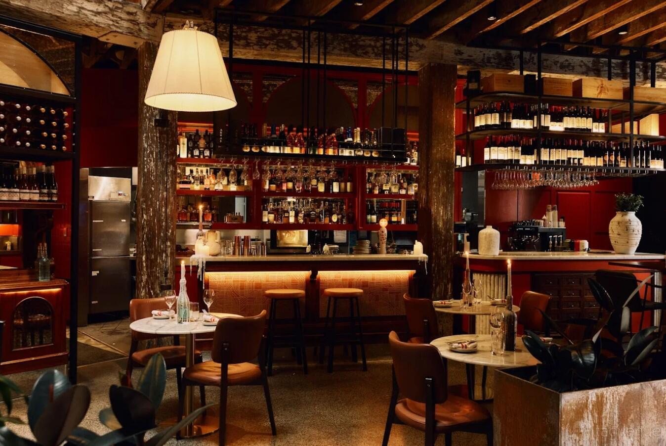 A restaurant with exposed wooden beams, a stocked backlit bar, and round tables with leather chairs at PERCY.