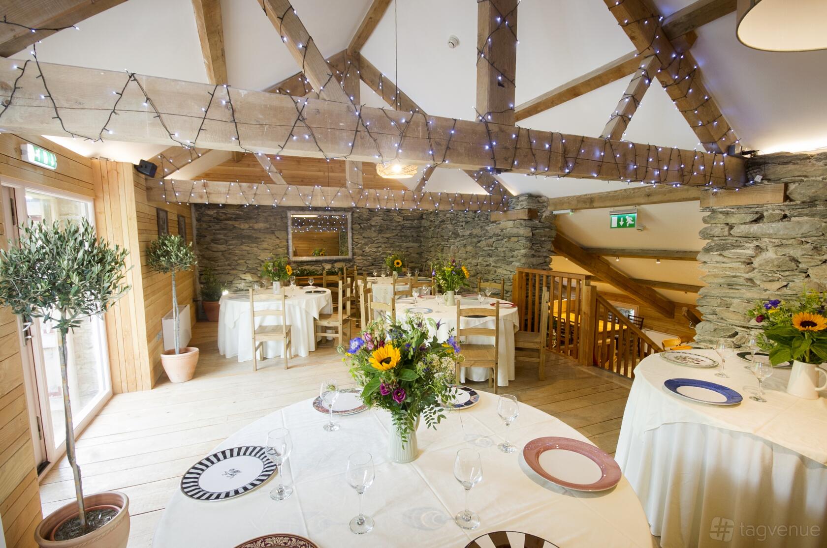 An event space in a barn with exposed wooden beams, stone walls, round tables, and string lights at Grove Barn.