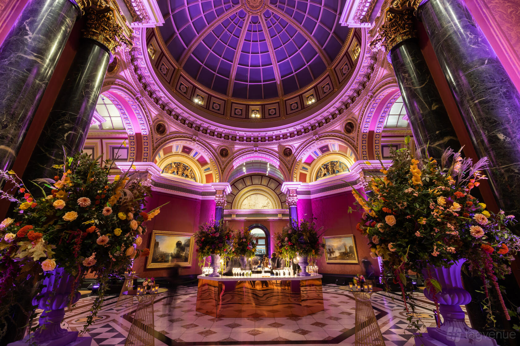 An event space with a domed ceiling, arched windows, uplighting, and large floral arrangements at The National Gallery.