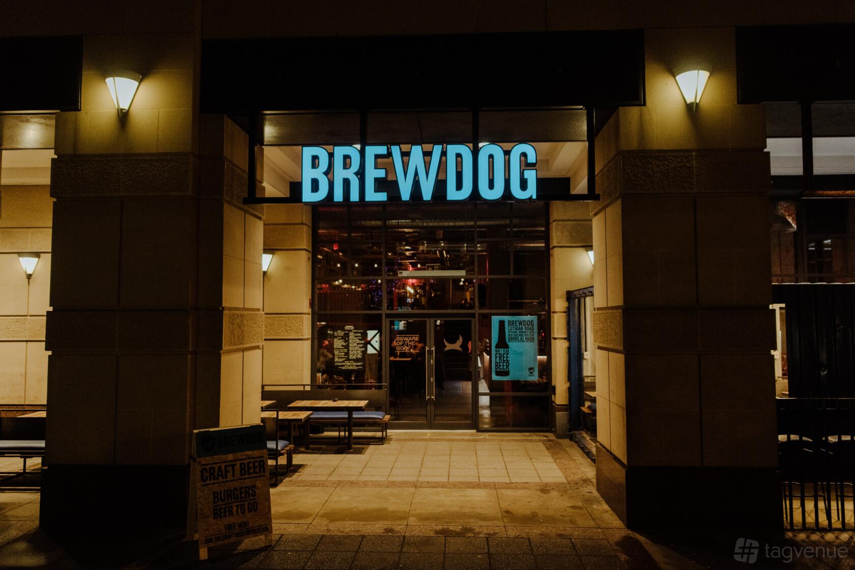 An entrance to a pub with large glass doors, illuminated signage, and outdoor benches at BrewDog Edinburgh Lothian Road.