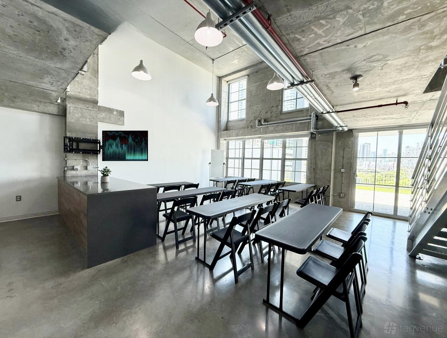 A photo studio with floor-to-ceiling windows, concrete walls, and rows of black tables and chairs at Crysp Studio.